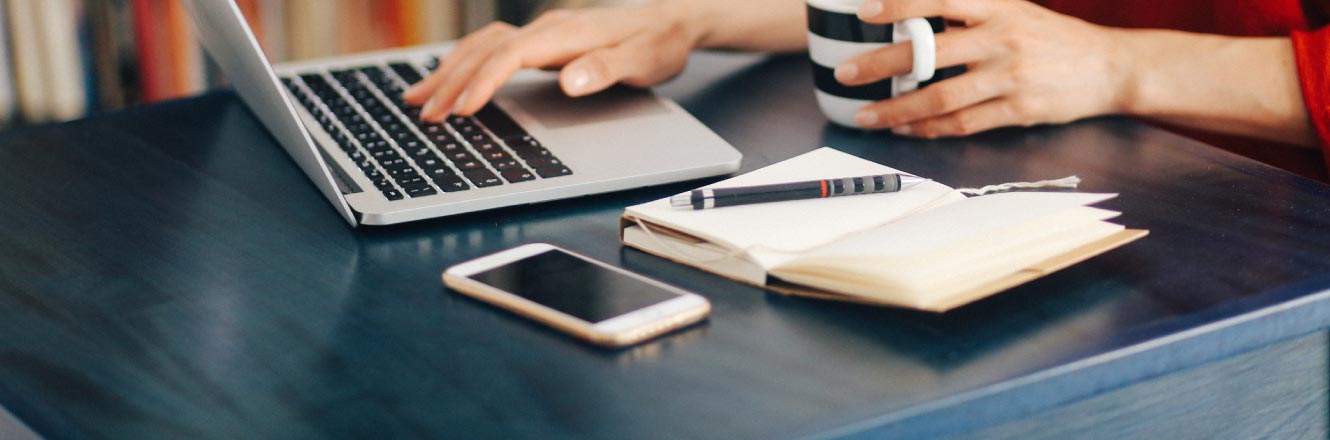 A woman having coffee and writing at a desk with a laptop and phone.