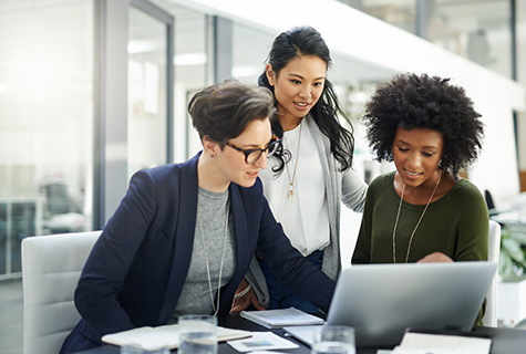 Three women review work together on computer in a bright office building